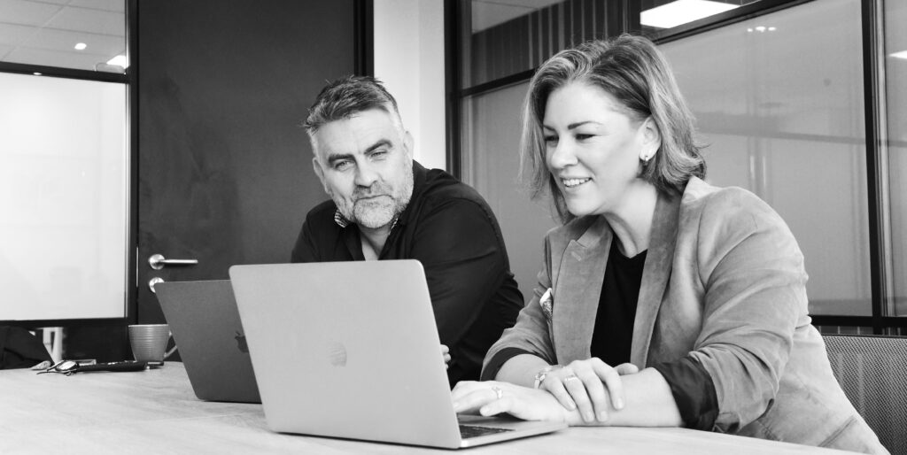 Two colleagues seated at a meeting table reviewing work on laptops in a glass‑walled office workspace.