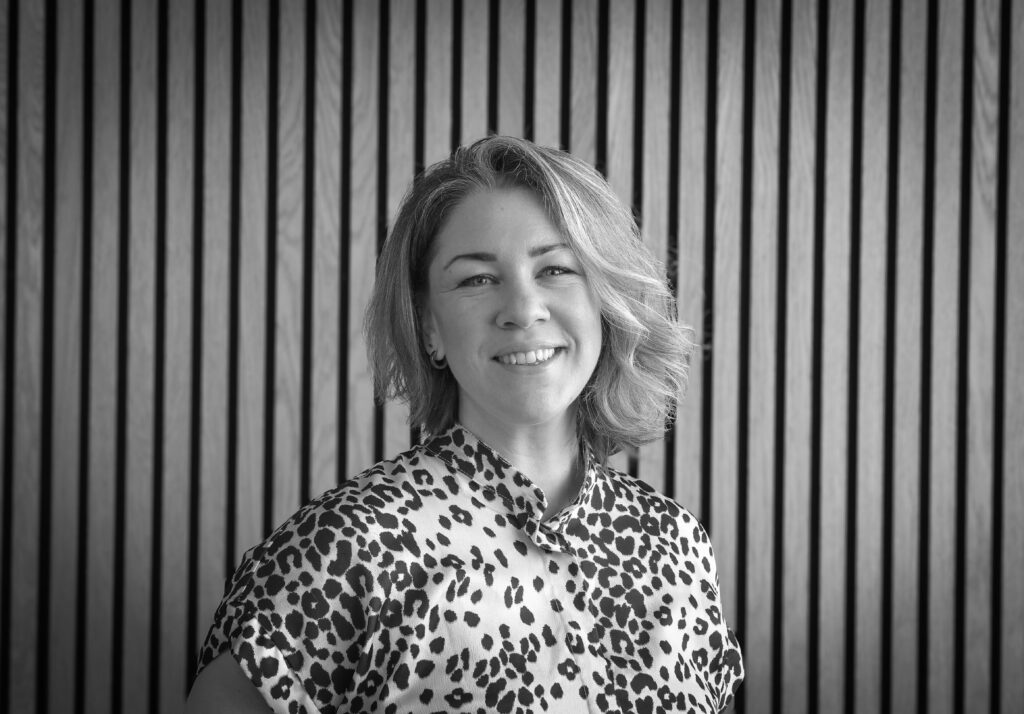 Black and white image of professional portrait of a person standing against a wood‑slat wall, wearing a patterned shirt in an office setting.