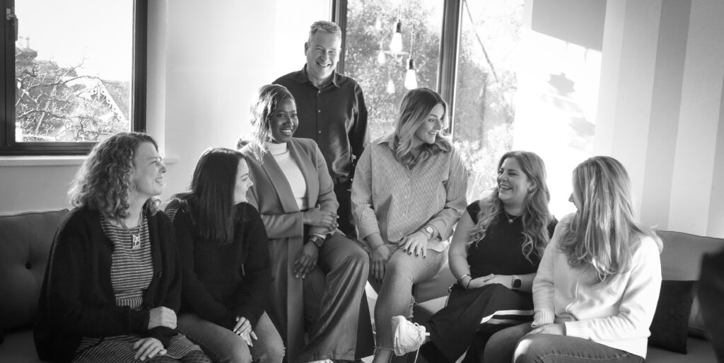 Group of colleagues seated and standing together in a bright office lounge, representing team culture and collaboration.