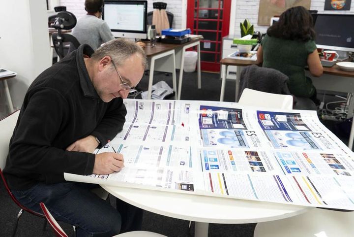 Person reviewing and marking up a large printed layout at a table, with colleagues working at desks in an open office.